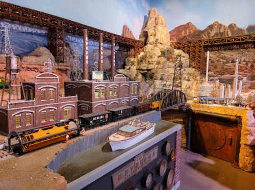 A detailed model train setup featuring buildings, a bridge, and a boat on water, set against a rocky landscape.