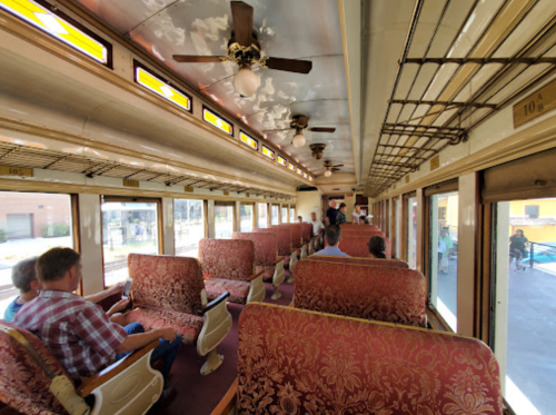 Interior of a vintage train car with patterned seats, ceiling fans, and passengers seated along the aisle.