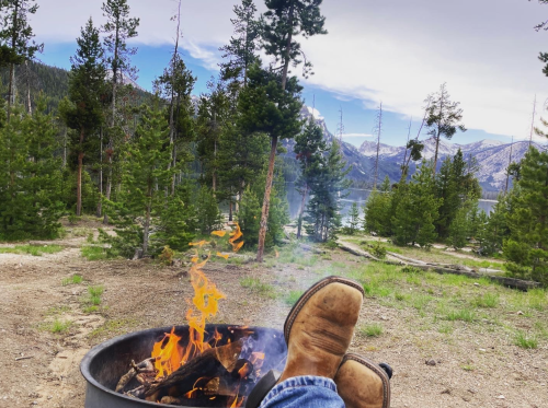 A person’s feet in boots resting near a campfire, surrounded by trees and mountains by a lake.