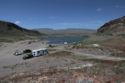 A scenic view of a lake surrounded by hills, with parked vehicles and RVs along the shore.