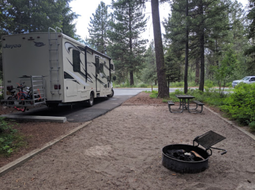 A parked RV in a wooded campsite with a picnic table and a fire pit in the foreground.
