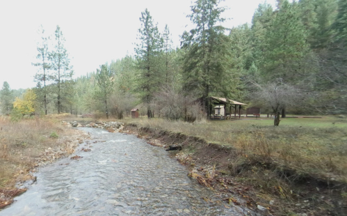 A serene stream flows beside a forested area with a cabin and picnic shelter in the background.