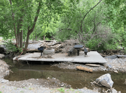 A concrete picnic area with grills, surrounded by trees and a shallow stream, showing signs of recent flooding.