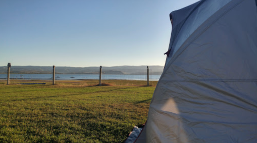 A tent set up on grassy land with a view of a calm lake and distant mountains under a clear blue sky.