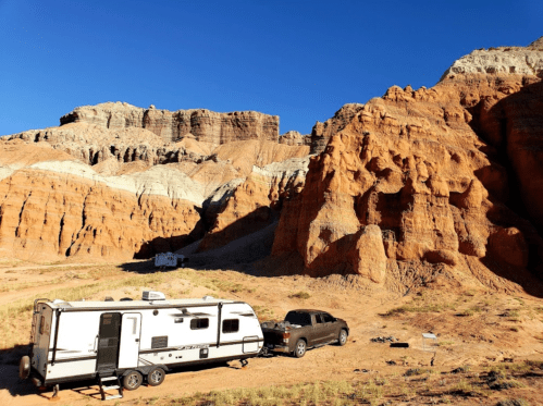 A camper trailer parked in a desert landscape with colorful rock formations and a clear blue sky.