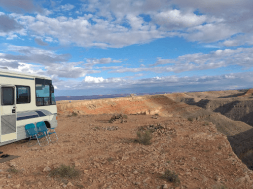 An RV parked on a rocky cliff with a chair nearby, overlooking a vast canyon under a partly cloudy sky.