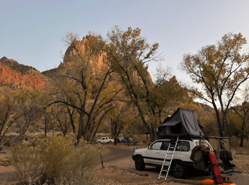 A campsite with a vehicle featuring a rooftop tent, surrounded by autumn trees and rocky cliffs at sunset.