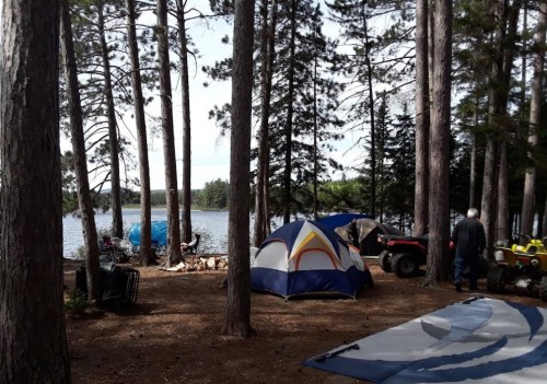 A campsite by a lake surrounded by tall trees, featuring tents, an ATV, and a peaceful outdoor setting.