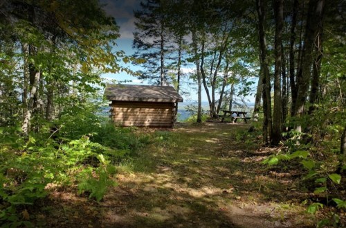 A wooden cabin surrounded by trees, with a picnic table nearby and a view of the landscape in the background.