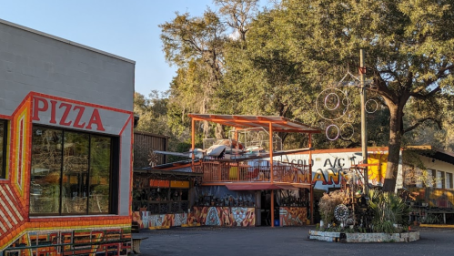 Colorful outdoor dining area with a pizza restaurant and unique art installations surrounded by trees.