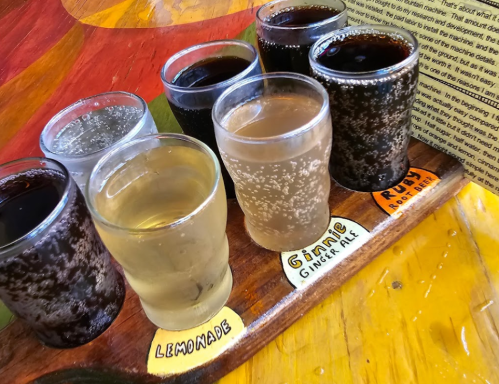 A wooden tray holds six small glasses of different beverages, including lemonade and ginger ale, on a colorful table.