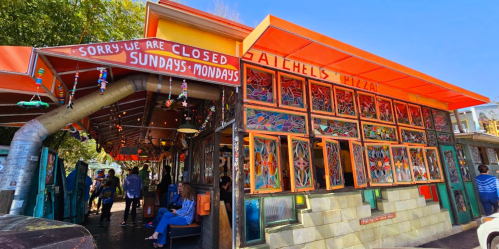 Colorful pizza shop with stained glass windows, outdoor seating, and a sign stating "Sorry, we are closed Sundays-Mondays."