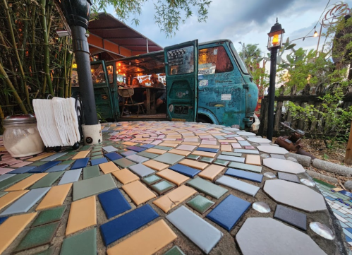 A colorful mosaic path leads to a vintage van in a cozy outdoor setting surrounded by greenery and warm lighting.