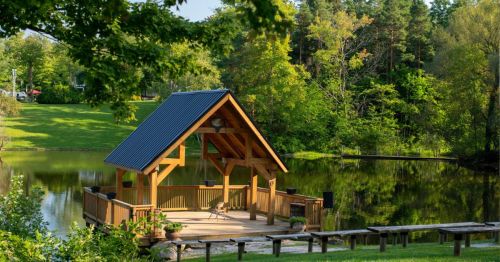 A wooden gazebo by a serene pond, surrounded by lush greenery and trees under a clear blue sky.
