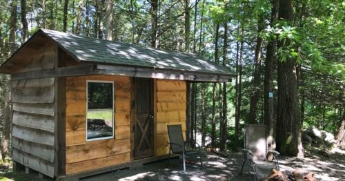 A small wooden cabin surrounded by trees, with two chairs outside and a picnic table in the background.