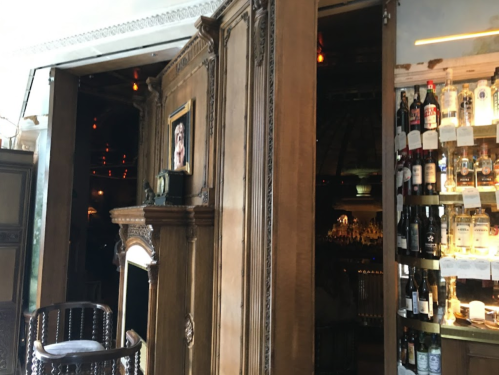 A wooden bar entrance with a framed picture, shelves of bottles, and a cozy, dimly lit atmosphere.