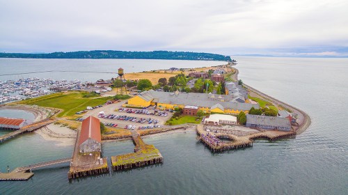 Aerial view of a coastal area with marinas, buildings, and a sandy shore under a cloudy sky.
