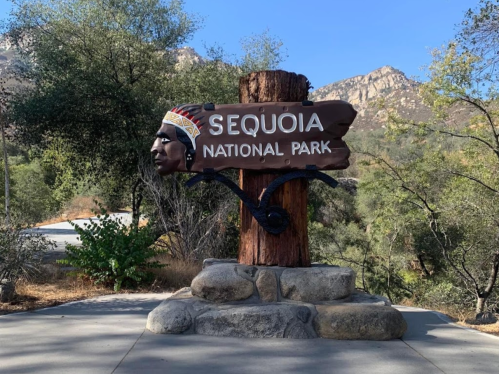 Sign for Sequoia National Park featuring a carved Native American face, set against a backdrop of trees and mountains.