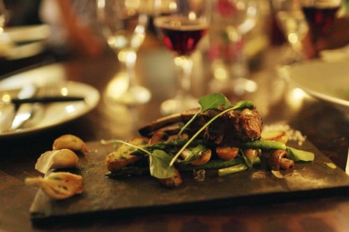 A beautifully plated dish of meat and vegetables on a slate, surrounded by wine glasses and plates on a wooden table.
