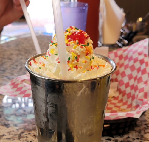 A metal cup filled with whipped cream, colorful sprinkles, and a cherry on top, sitting on a table.