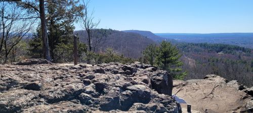 A rocky outcrop overlooks a scenic valley with distant hills under a clear blue sky.