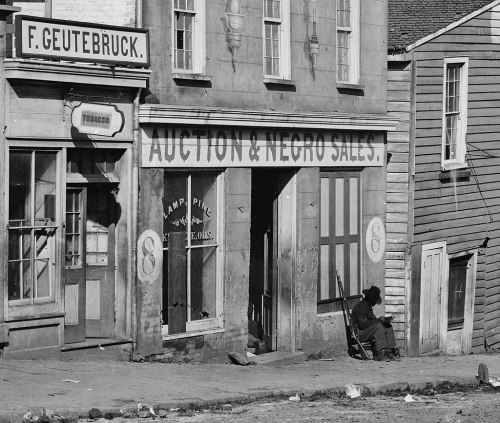 A historic black-and-white photo of a storefront with "Auction & Negro Sales" sign, featuring a person sitting outside.