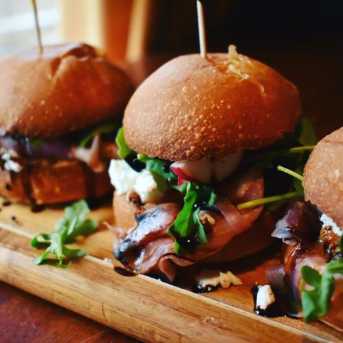 Three gourmet sliders on a wooden platter, featuring greens, cheese, and a drizzle of balsamic glaze.