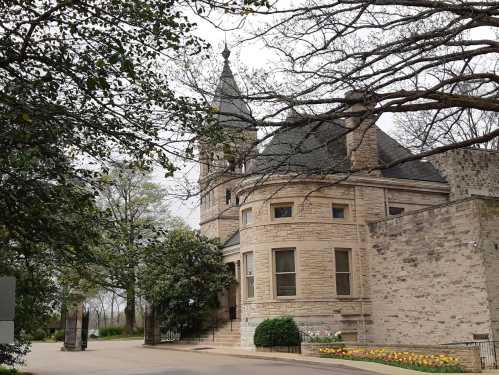 A stone building with a tower, surrounded by trees and colorful flowers, on a cloudy day.