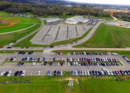 Aerial view of a large parking lot filled with cars, adjacent to a sports field and a building in a green landscape.