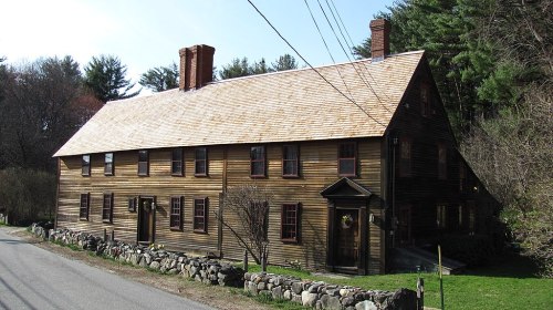 A historic wooden house with a sloped roof, surrounded by trees and a stone wall, set along a quiet road.