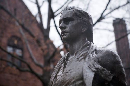 A close-up of a rain-soaked statue of a young man, with trees and a brick building blurred in the background.
