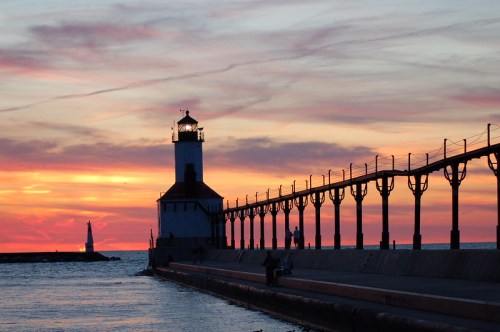 Silhouette of a lighthouse at sunset, with a pier extending into the water and colorful clouds in the sky.