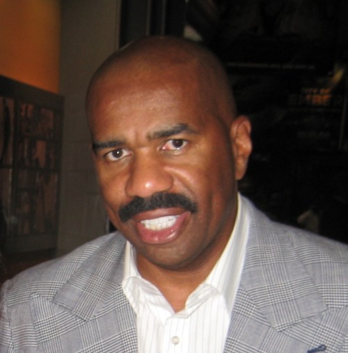 A man with a bald head and mustache, wearing a gray blazer, smiles while looking at the camera.