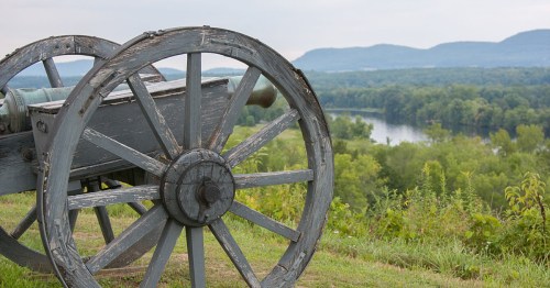 A weathered cannon on a grassy hill overlooking a river and distant mountains under a cloudy sky.