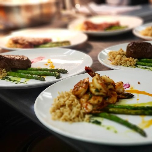Plates of gourmet food featuring shrimp, asparagus, rice, and a meat portion, arranged elegantly in a kitchen setting.
