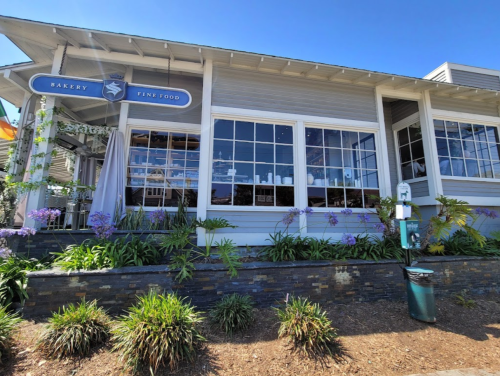 A charming bakery and fine food restaurant with large windows, surrounded by greenery and flowers on a sunny day.