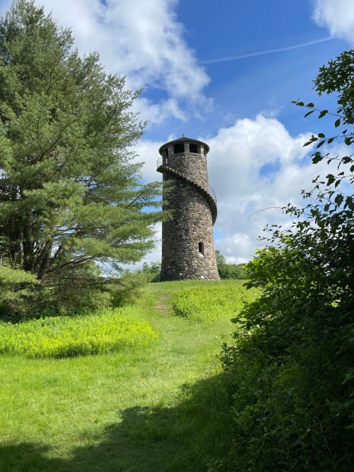 A stone tower with a spiral staircase stands on a grassy hill, surrounded by trees and blue skies.