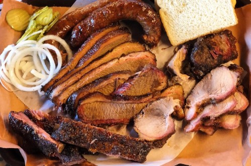 A platter of assorted smoked meats, including sausage, brisket, and ribs, served with pickles, onions, and bread.