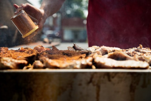 A person sprinkles seasoning on grilled meats sizzling on a barbecue.
