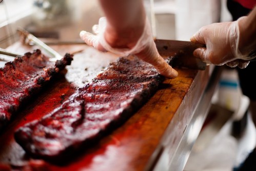 A person in gloves slices smoked ribs on a wooden cutting board, showcasing the juicy meat and charred edges.