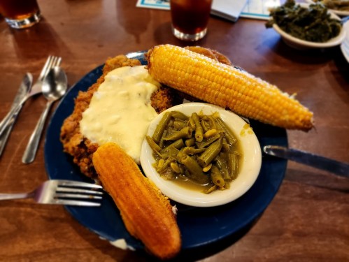 A plate of fried chicken with gravy, corn on the cob, and green beans, served with cornbread on a wooden table.