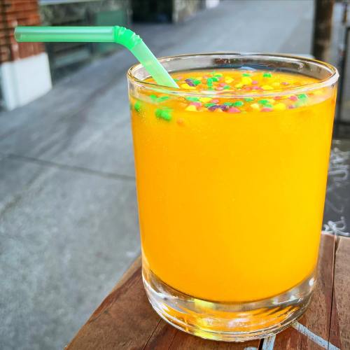 A vibrant orange drink with colorful sprinkles and a green straw, sitting on a wooden table outdoors.