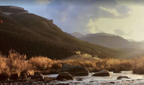 A serene landscape featuring a river, rocky foreground, and mountains under a cloudy sky with autumn foliage.