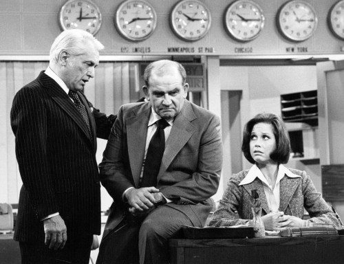 Three characters in a newsroom setting: two men in suits and a woman in a blouse, engaged in a serious discussion.