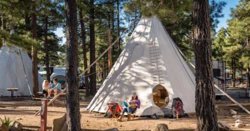 A white teepee surrounded by trees, with people relaxing nearby in a campsite setting.
