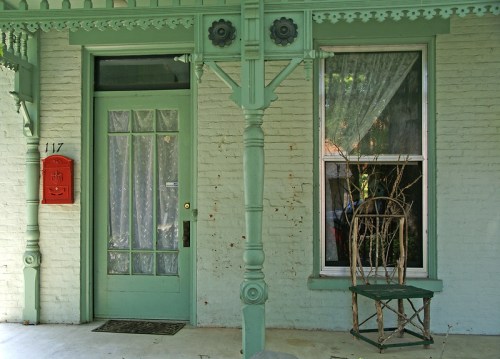 A green door with a decorative frame, a red mailbox, and a window with sheer curtains beside a bare branch in a chair.