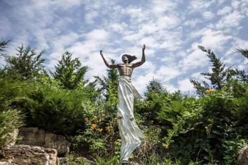 A tall statue of a figure with outstretched arms, surrounded by greenery and a blue sky with clouds.