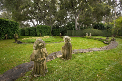 A serene garden with stone sculptures, lush greenery, and a winding path surrounded by hedges and trees.