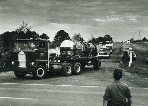 A black and white photo of a truck carrying large tanks, with a person watching from the roadside.