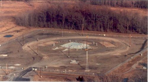 Aerial view of a large, fenced industrial site with machinery and structures, surrounded by trees and open land.
